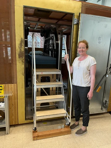 Woman poses smiling next to a set of portable stairs that lead up to a platform for an experimental MRI setup. 