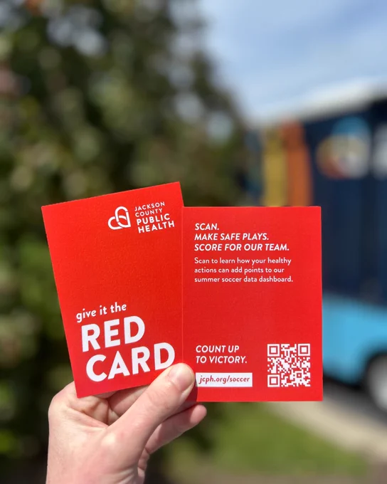 A hand holds two red cards featuring the Jackson County Public Health logo. The text on the cards encourages scanning to learn how healthy actions can contribute to a soccer data dashboard. A QR code and a web address "jcph.org/soccer" are also visible. The background is slightly blurred, showing greenery and a hint of a colorful mobile health truck.
