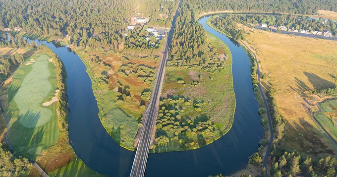 Aerial view of a bridge crossing a river that splits and forms an island. The landscape includes lush greenery, wooded areas, and a golf course on one side. An expanse of grassland is visible on the other side.