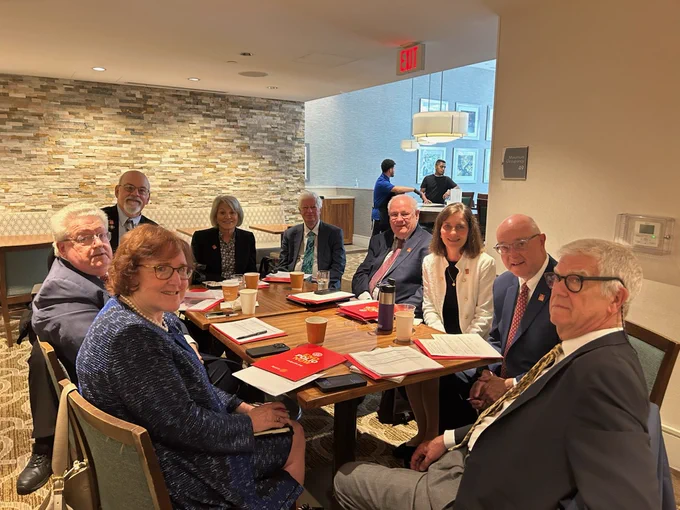 A group of people are seated around a table covered with coffee cups, folders, and notepads. The setting appears to be a meeting or conference room with a neutral-toned interior and a stone accent wall.