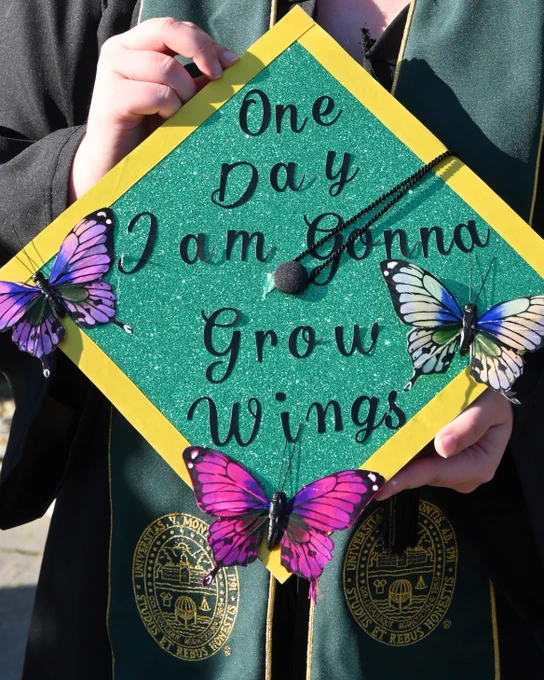 A person holds a decorated graduation cap featuring colorful butterflies and the quote "One Day I Am Gonna Grow Wings." The cap has a sparkly green background and yellow border. The person is wearing a black gown with patches bearing the seal of University Of Vermont.