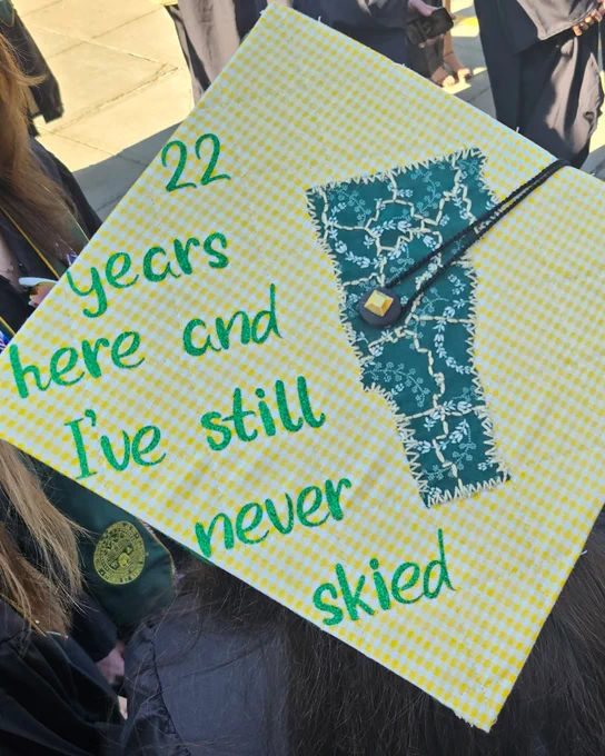 A graduation cap with yellow and white checkered fabric features a green cutout of Vermont and the words "22 years here and I've still never skied" written in green.