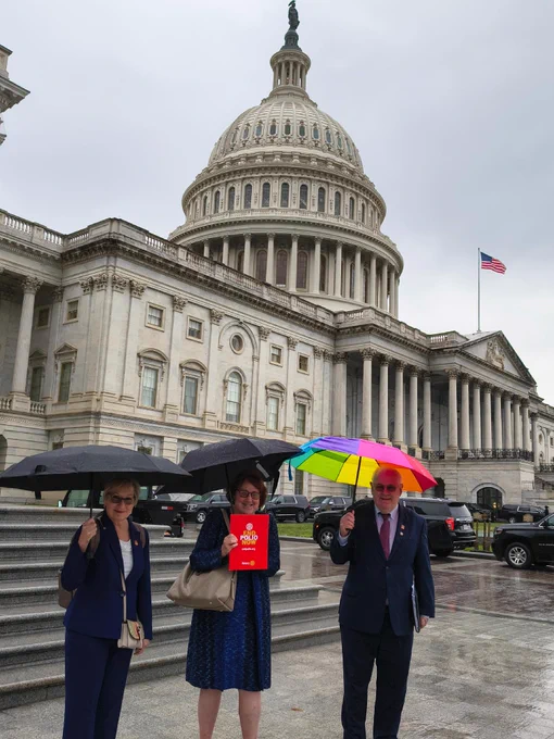 Three individuals holding umbrellas stand in front of the United States Capitol building on a rainy day. One person holds a brightly colored umbrella and another person holds a red folder.