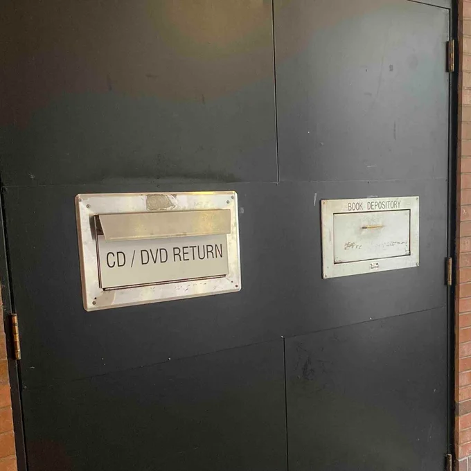 Black wall with two metal return slots labeled CD/DVD RETURN and BOOK DEPOSITORY for media and book returns.