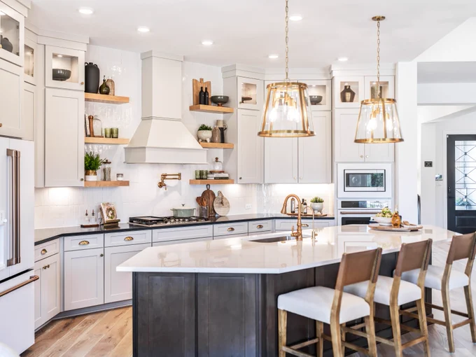 A stylish, modern kitchen by David Weekley Homes featuring light cabinets and a large kitchen island with a dark finish and four wooden chairs. Hanging pendant lights provide illumination. The space includes a built-in oven, a refrigerator, and open shelving with various decorative items. The room has natural wood flooring and recessed ceiling lights, creating a bright and inviting atmosphere.