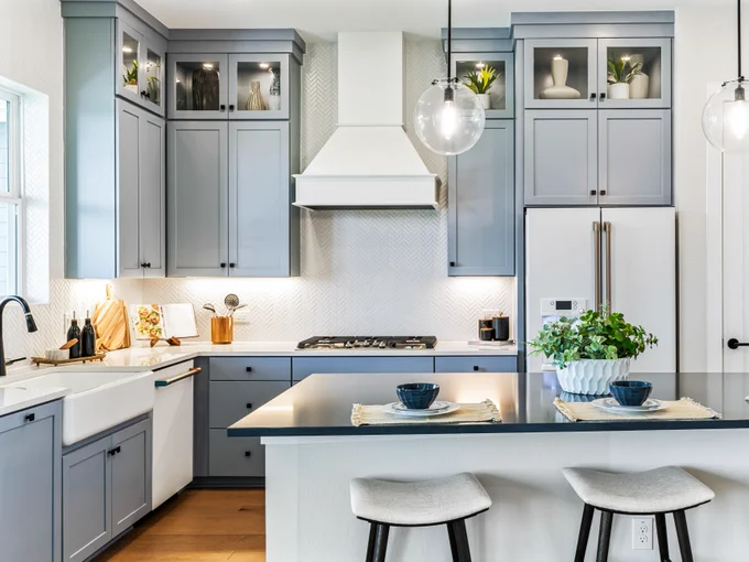 A modern kitchen by David Weekley Homes with light blue cabinets and a white backsplash. The room features a farmhouse sink, a gas cooktop, and a white refrigerator. Three bar stools are placed at the kitchen island, which is set with two place settings. Two spherical pendant lights hang above the island. Small plants and decor items are on the upper cabinets.
