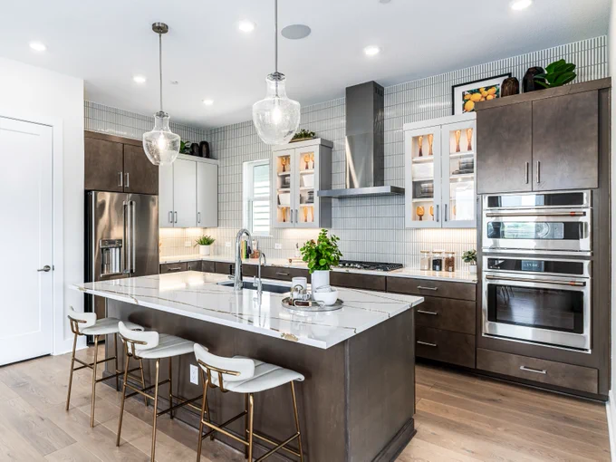 A modern kitchen by David Weekley Homes with a large island, featuring white countertops and four stools. There are stainless steel appliances, including a fridge and double oven. Open shelving displays plants and dishes. Pendant lights hang above the island, and light wood flooring adds warmth.