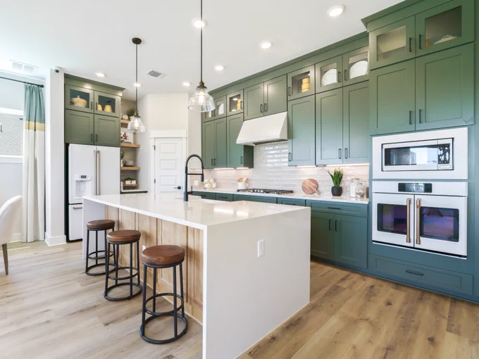 A modern kitchen by David Weekley Homes with green cabinetry and stainless steel appliances. It features a large white island with wooden bar stools. The backsplash is white subway tile, and the flooring is light wood. To the left is a small dining area with a table and chairs near a window. Pendant lights hang from the ceiling.