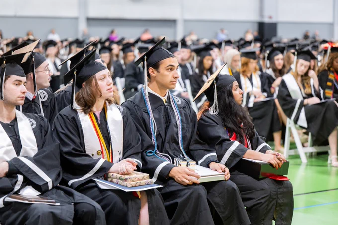 Graduates seated in rows wearing black caps and gowns with white stoles and different colored cords, attentively watching a ceremony. The setting appears to be an indoor graduation event.