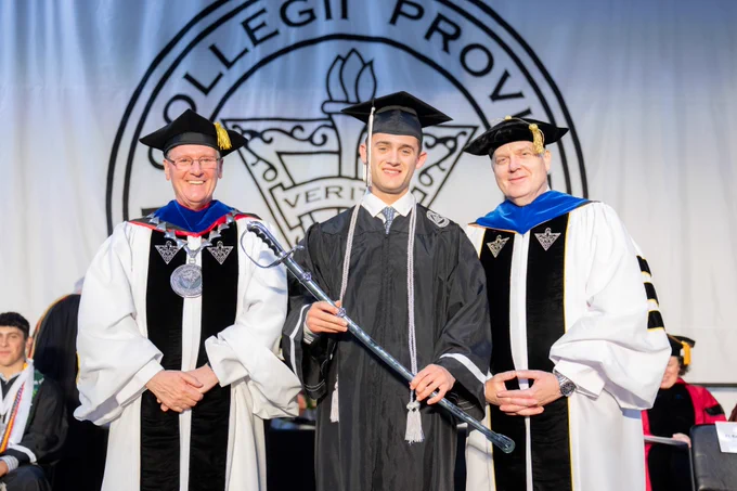 Three people in academic regalia stand in front of a Collegii Prov insignia. One in the center holds a ceremonial mace.