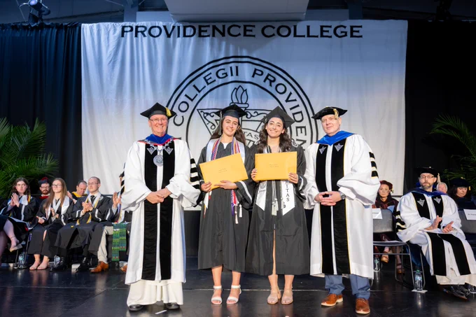 A graduation ceremony at Providence College. Four individuals stand on stage, two in academic regalia holding diplomas, flanked by two others in official robes. The background features a large Providence College banner and several seated individuals in similar attire.