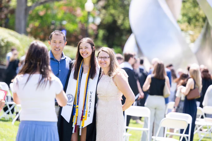 A graduate in cap and gown poses outdoors with two people. The group is smiling, and it appears to be a sunny day.