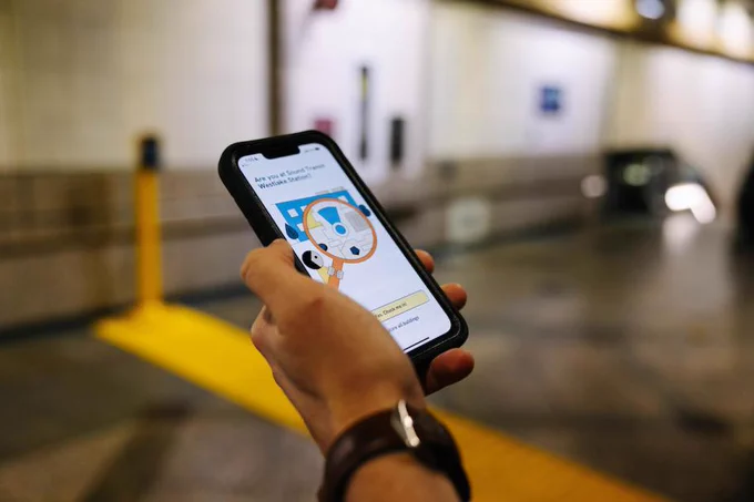 A person holds a smartphone displaying a page from the GoodMaps app. The background shows an indoor station setting with blurred architectural details.