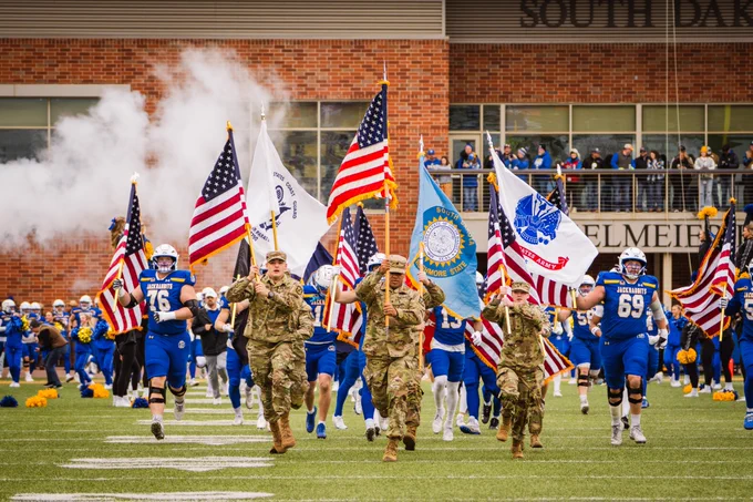 Members of a military color guard in camouflage uniforms run onto a football field carrying multiple flags, including the United States flag, a South Dakota state flag, and military branch flags, while SDSU football players in blue uniforms run behind them through smoke near the stadium entrance, with spectators watching from a brick concourse and seating areas in the background.