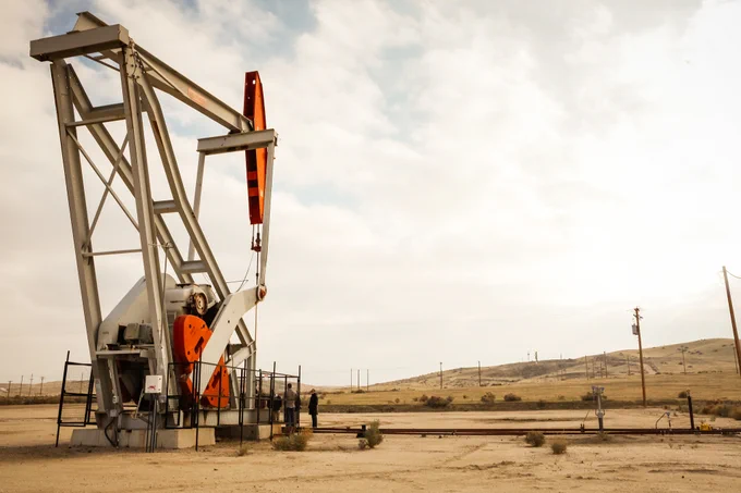 Oil pumpjack operating in a dry, open landscape under a cloudy sky with distant hills and utility poles.