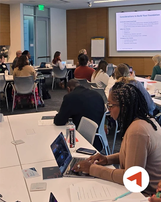 A group of people sitting at tables in a conference room, attentively listening to a presentation. A screen displays a slide titled "Considerations to Build Your Foundation." The Ascendium logo is in the lower right corner.
