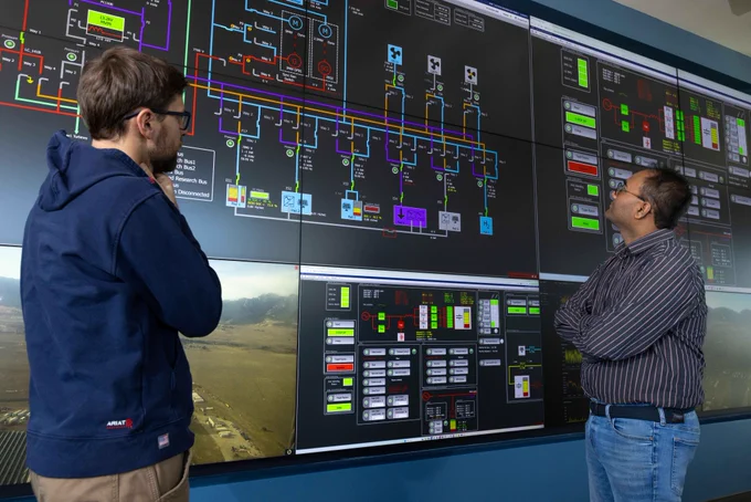 Two individuals are observing a large wall-mounted digital display featuring complex technical diagrams and data related to a power grid system. The screen includes various boxes, lines, and colored indicators.