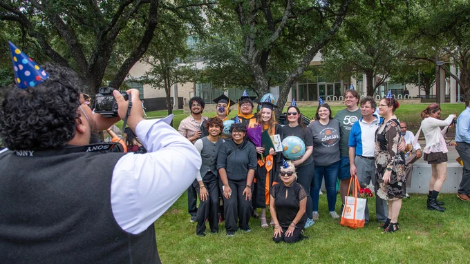 A group of people, some wearing graduation caps, pose for a photo outdoors on a grassy area. One person is taking the photo. In the background, there's a large building and several trees.