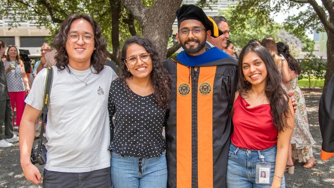 A group of four people stand together outdoors, smiling at the camera. One person is wearing graduation attire with an orange and black gown and a cap. Trees and other people are visible in the background.