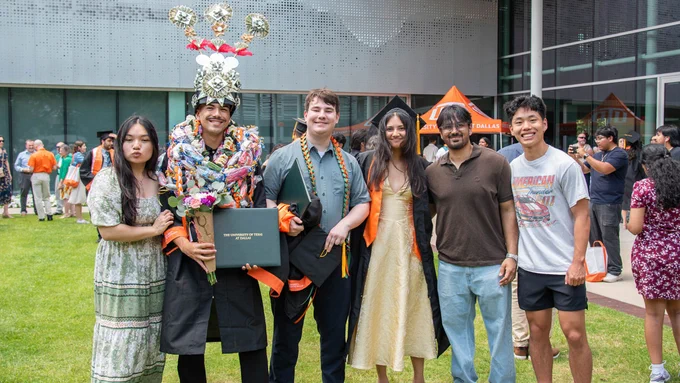 A group of people is celebrating graduation outside the University of Texas at Dallas. One person is wearing a colorful decorative garland and holding a diploma. Others are smiling, and one holds a bouquet of flowers.