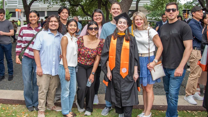 A group of people smiling and standing together outdoors. One person in the center is wearing a graduation cap and gown with orange stoles. Trees and a building are visible in the background.