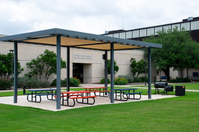 A covered outdoor seating area with colorful picnic tables is situated on a lawn. Trees and plants surround the building. A barbecue grill and a small trash bin are nearby.