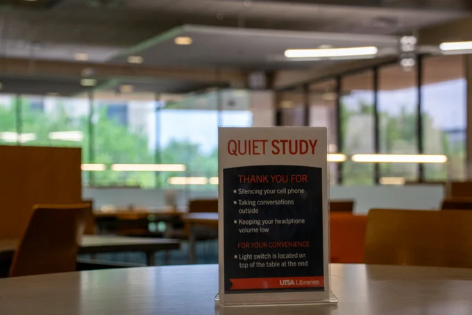 A sign on a table in a library reads "Quiet Study" with guidelines for maintaining silence. The background shows a well-lit room with tables and chairs, and large windows revealing outdoor scenery. The sign mentions UTSA Libraries.