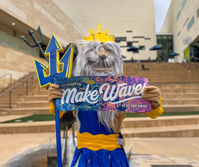 King Triton holds a sign reading "I'm ready to make waves at UC San Diego" while standing in an outdoor area with steps and modern buildings in the background.