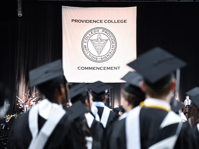 Graduates in caps and gowns face a large "Providence College Commencement" banner. The college seal is visible on the banner.