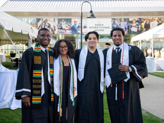 A group of four graduates in caps and gowns pose together outdoors at a graduation ceremony. They wear stoles and cords. A building with a banner saying "Seek Something More" is visible in the background.