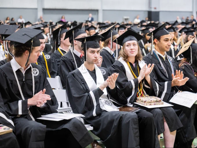 A group of graduates in caps and gowns sitting in rows, clapping during a commencement ceremony.