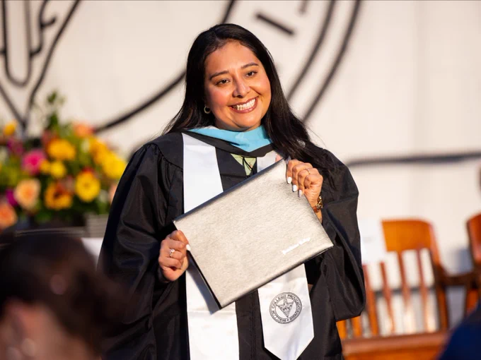 A person in graduation attire holds a diploma folder while smiling. A logo is visible on the folder. In the background, there are colorful flowers and several chairs.