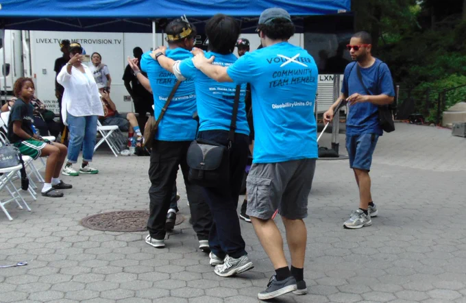 Three volunteers in bright blue Disability Unite t-shirts form a conga line, each with hands on the shoulders of the person ahead, with a crowd of festival attendees gathered in the sunny park behind them.