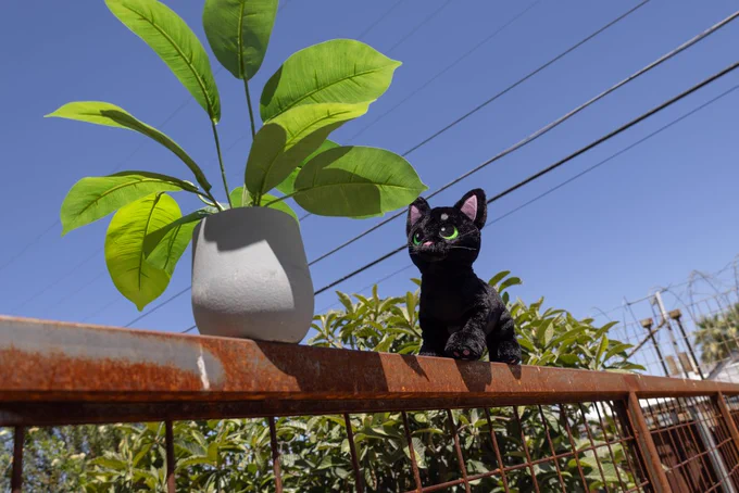 A photo of the Little Kitty plush outdoors on a railing, looking like it's about to knock over a worryingly precarious potted plant. 