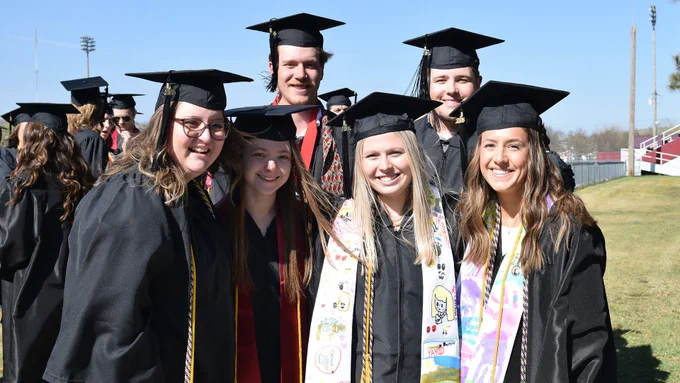 group of 6 graduates outdoors, smiling at camera with other graduates in the background