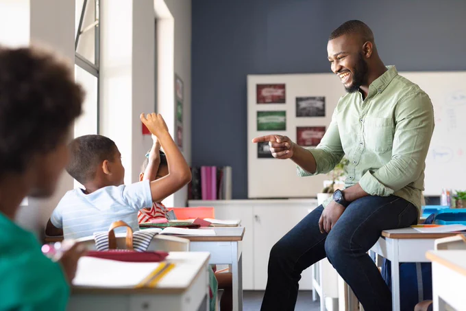 A male teacher in a classroom points to a young student whose hand is raised. 
