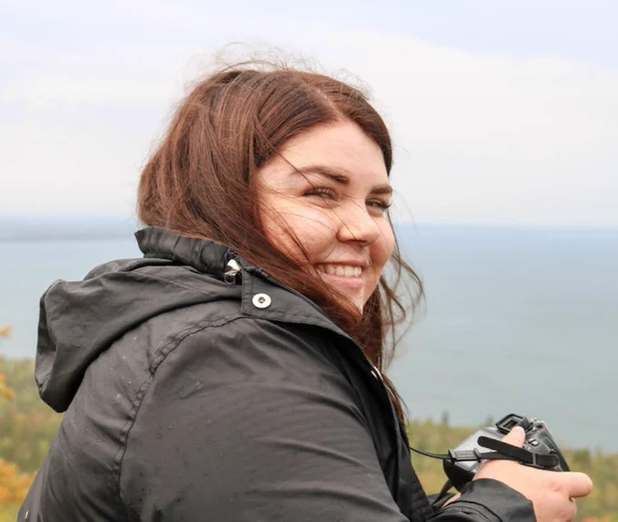 Person in a black rain jacket holding a camera, overlooking a calm sea with a cloudy sky.
