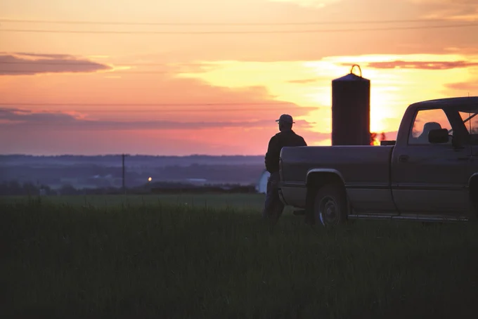 Silhouette of a person standing beside a pickup truck in a field during a vibrant sunset with orange and purple hues.