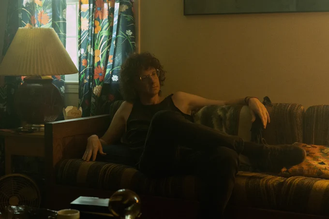 From Widow’s Bay. Chris Fleming as Tod the Shaman sits in a lounged position on a couch in dim lighting with his legs crossed.
