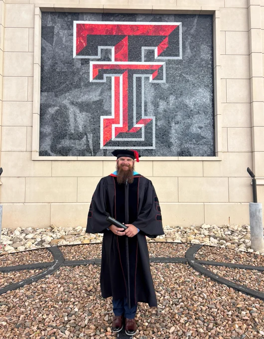 A person in graduation attire stands holding a diploma in front of a large Texas Tech University logo on a wall with a rocky ground in front.