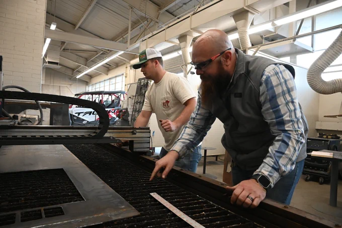 Two people operate equipment in an industrial setting. One adjusts a metal sheet, while another observes. The workshop is spacious with machinery and ventilation pipes.