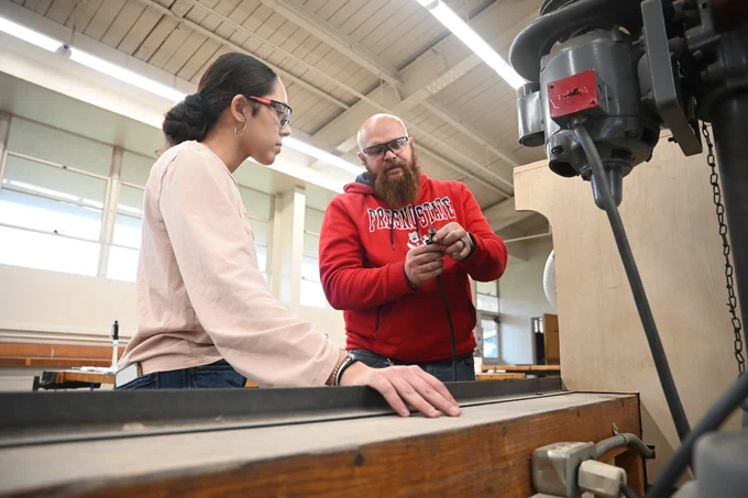 A person in a red Fresno State hoodie explains a tool to another person in a workshop setting. Both are wearing safety glasses. The room has high ceilings with large windows.