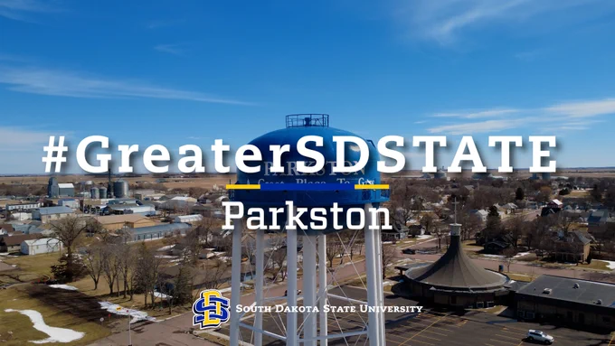 A water tower stands prominently in Parkston, featuring the text "#GreaterSDSTATE" and "South Dakota State University" with the university's logo. The surrounding area includes trees and buildings under a clear blue sky.