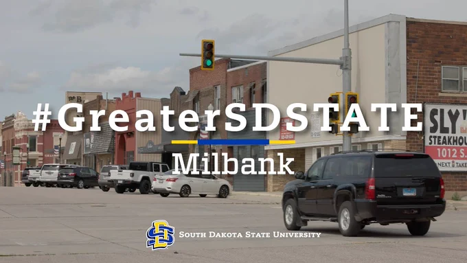 Street view of Milbank with several buildings and parked vehicles. A sign for Sly's Steakhouse is visible. Text overlay reads "#GreaterSDSTATE Milbank" with the South Dakota State University logo.