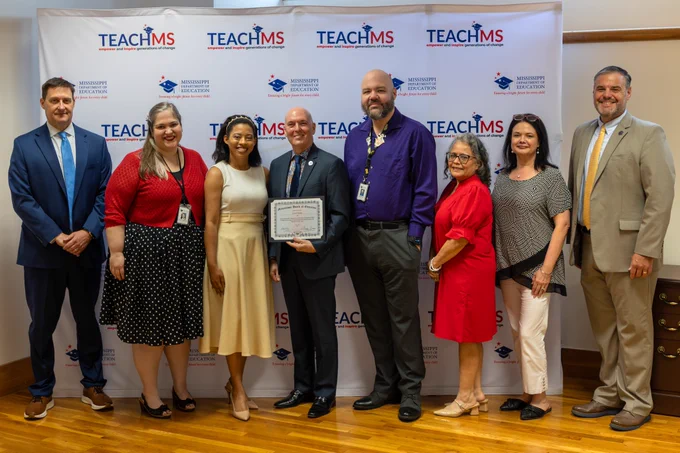This image shows eight people standing side by side with the person in the middle holding a certificate. 