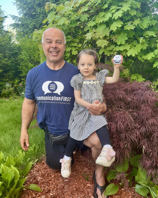 [Image 1: A grandfather wearing a CommunicationFIRST shirt bends down on one knee and holds his granddaughter who is a toddler with a CommunicationFIRST button in her hand. The two of them are in a yard with lush trees.]