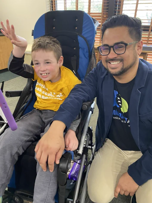 [Image 2: A young AAC user sits with his AAC device mounted in front of him, and he happily waves. His smiling dad squats down beside him. The pair wear CommunicationFIRST shirts and pose in front of windows letting in natural light.]