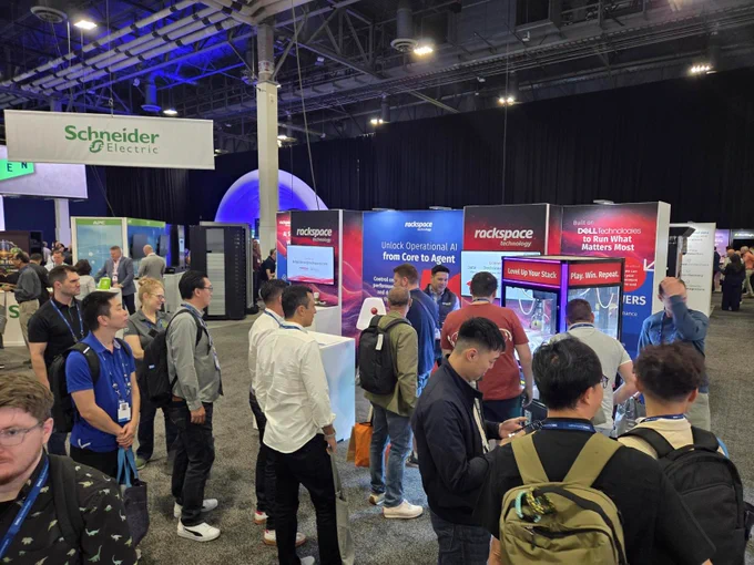 People are gathered in a convention center with booths from Schneider Electric and Rackspace. Attendees are engaged with displays and conversations.