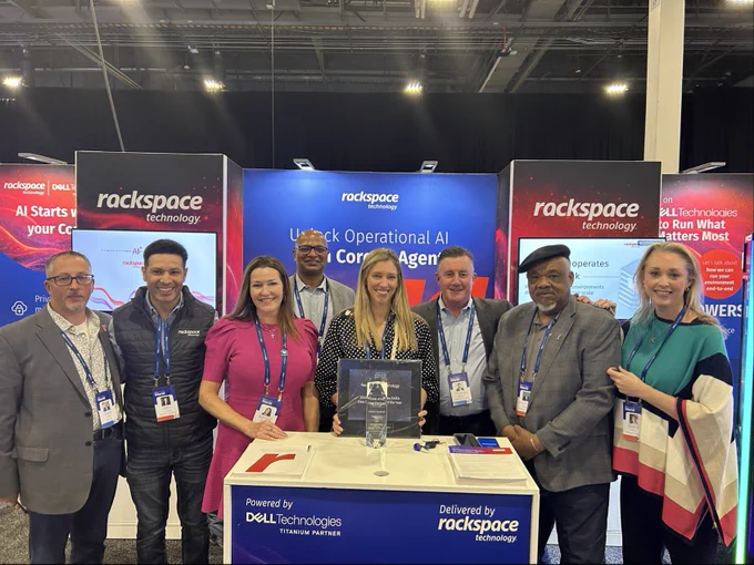 A group of eight individuals standing together at a tech conference booth featuring Rackspace Technology and Dell Technologies branding. They're smiling at the camera, with some wearing conference badges. The booth displays promotional materials related to AI and cloud services.