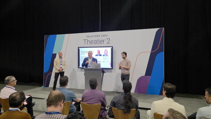 A group of individuals is gathered in front of a presentation area labeled "Solutions Expo Theater 2." One person speaks at a podium, while another stands nearby. An audience is seated, attentively listening. The setup includes a screen displaying content related to the event.